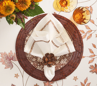 A table setting featuring a floral embroidered tablecloth, brown woven placemat, patterned dinner plate, folded white napkin with brown trim held by a Kim Seybert Begonia Napkin Ring Set of 4, two amber glasses, and a bouquet of orange flowers.