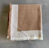 An Eyelash Alpaca Throw by Alicia Peru Home, featuring a light brown hue with a white border and fringe, is folded and placed on a gray surface.