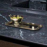 The Anna New York Striations Gold Nut Bowl, with two spoons and green olives, rests on a matching gold tray atop a black marble counter—an eye-catching modern display.