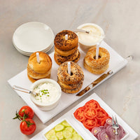 Bagels are artfully arranged on the Inspired Generations Acrylic Bagels On Board Tray - White, accompanied by cream cheese topped with serving knives and surrounded by sliced tomatoes, cucumbers, and red onions. Stacked plates in the background complete this perfect brunch board setup.