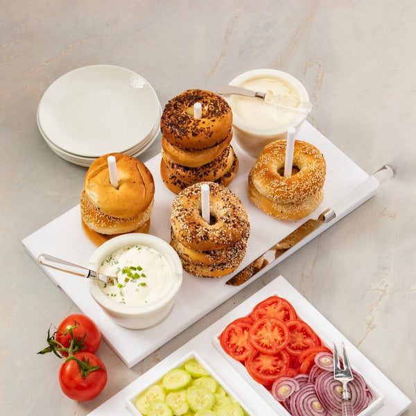 Bagels are artfully arranged on the Inspired Generations Acrylic Bagels On Board Tray - White, accompanied by cream cheese topped with serving knives and surrounded by sliced tomatoes, cucumbers, and red onions. Stacked plates in the background complete this perfect brunch board setup.