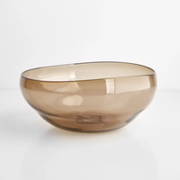 A translucent, light brown handblown bowl from the Gary Bodker Nesting Glass Bowl Collection Wheat sits on a white surface against a plain background—an elegant piece from this stackable set.