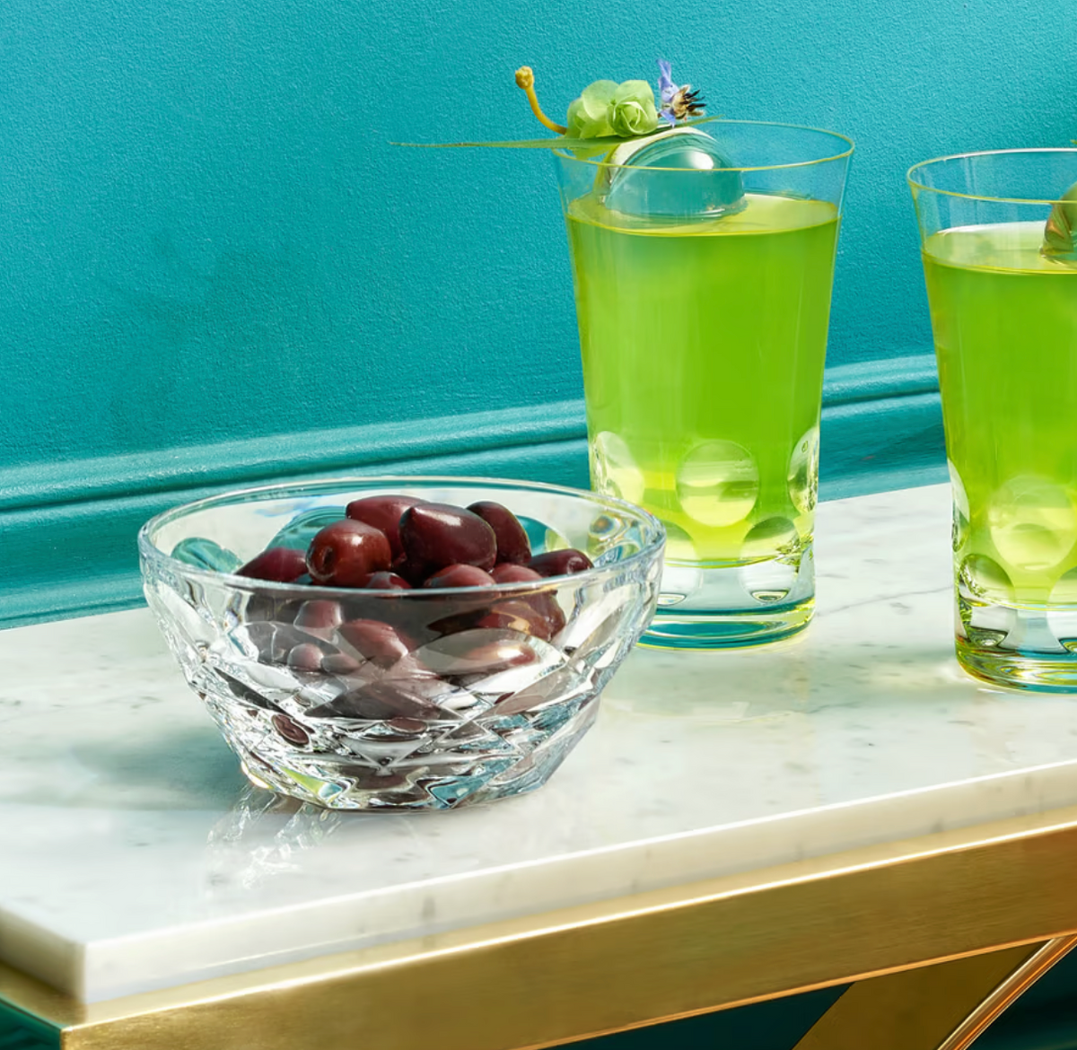 A Baccarat Swing Bowl filled with dark olives sits on a marble table beside two tall glasses of green drinks with floral garnishes and ice spheres, set against a turquoise wall for a stylish brunch display.