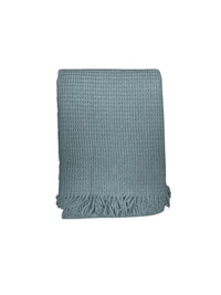 The Basic Waffle Throw by A Soft Idea is a light blue cotton acrylic blanket with a textured waffle weave and fringed edges. Machine washable for easy care, it combines style and comfort, shown folded against a white background.