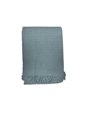 The Basic Waffle Throw by A Soft Idea is a light blue cotton acrylic blanket with a textured waffle weave and fringed edges. Machine washable for easy care, it combines style and comfort, shown folded against a white background.