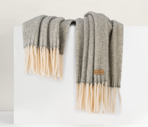 The Lands Downunder Andes Throw, a cozy grey alpaca wool blanket with cream fringe, is draped over a white surface.
