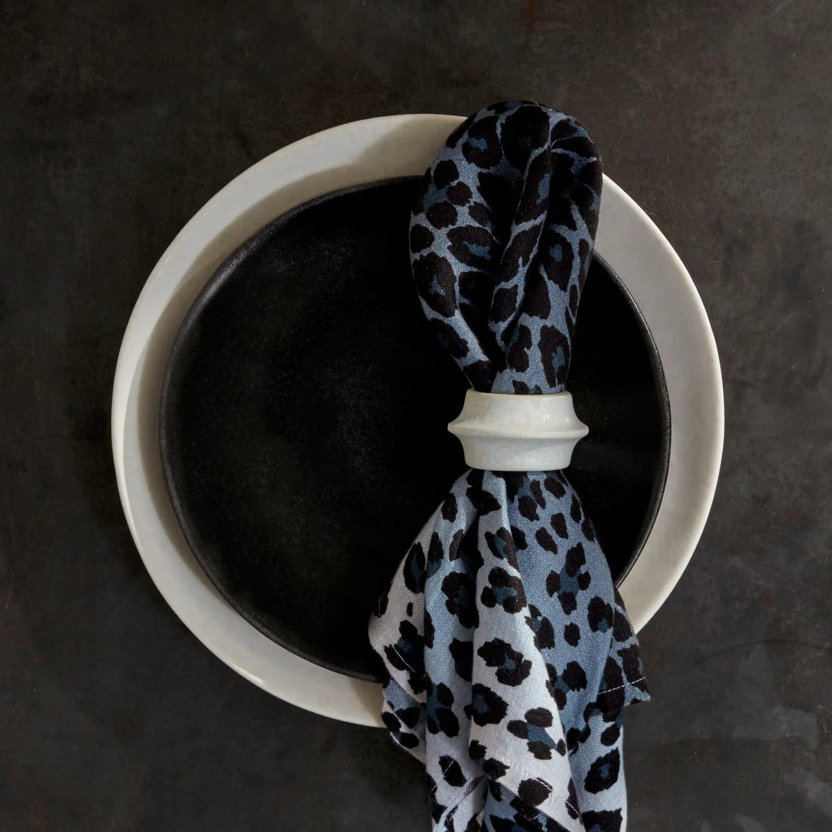 A blue and black leopard-print napkin with a L'Objet Terra Cuff Napkin Ring rests atop stacked black and white plates on a dark surface.