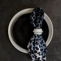 A blue and black leopard-print napkin with a L'Objet Terra Cuff Napkin Ring rests atop stacked black and white plates on a dark surface.