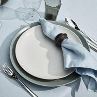 A close-up of a table setting featuring a white plate, gray charger, fork, knife, spoon, glass, tinted tumbler on a light blue tablecloth and a light blue napkin styled with the L'Objet Terra Cuff Napkin Ring Set of 4.