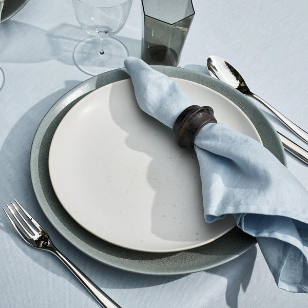 A close-up of a table setting featuring a white plate, gray charger, fork, knife, spoon, glass, tinted tumbler on a light blue tablecloth and a light blue napkin styled with the L'Objet Terra Cuff Napkin Ring Set of 4.