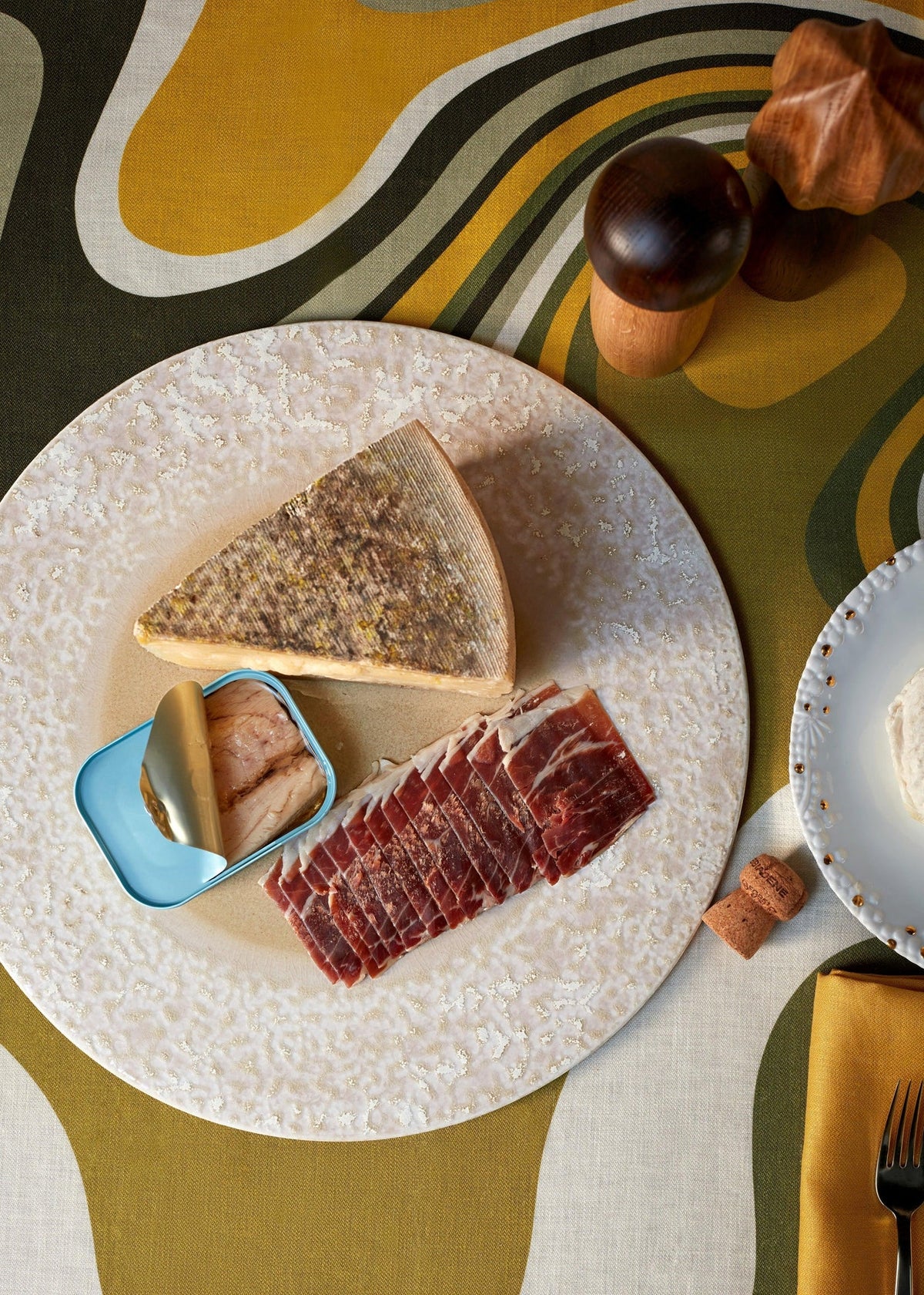 On the Terra Platter Charger Dusty Rose by L'Objet, a wedge of cheese, tinned fish, and sliced cured meat are arranged atop retro porcelain placemats, with salt and pepper shakers, a cork, and part of another plate nearby.