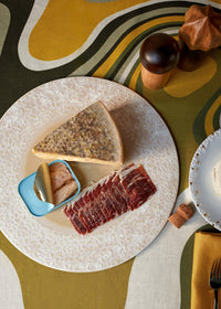 On the Terra Platter Charger Dusty Rose by L'Objet, a wedge of cheese, tinned fish, and sliced cured meat are arranged atop retro porcelain placemats, with salt and pepper shakers, a cork, and part of another plate nearby.
