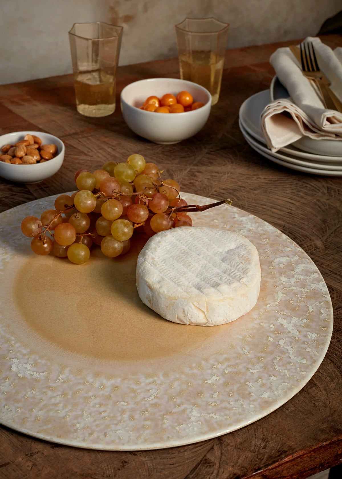 A round wheel of soft white cheese and green grapes sit on a L'Objet Terra Platter Charger in Dusty Rose, atop a wooden table surrounded by bowls of snacks, stacked plates, napkins, and glasses.
