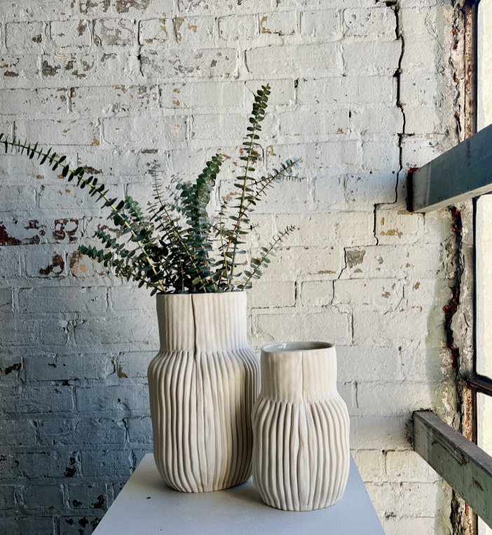 Two CYM Warkov Ceramics Series 02 Bottle Vases in cream sit on a white surface before a brick wall. One hand-sculpted porcelain vase displays green eucalyptus stems, while the other remains empty as sunlight streams in from the right.