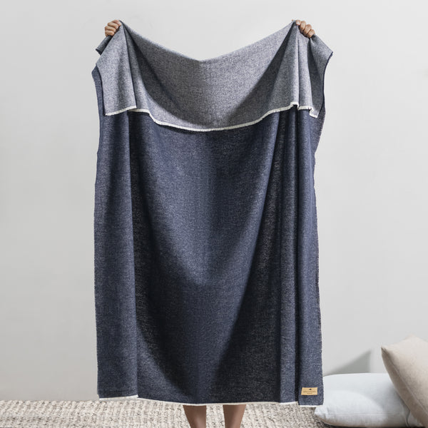 Someone holds up the Aspen Reversible Throw by Lands Downunder in blue and gray, its soft, cozy texture covering them. The indoor setting features a light wall and a textured rug on the floor.