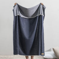 Someone holds up the Aspen Reversible Throw by Lands Downunder in blue and gray, its soft, cozy texture covering them. The indoor setting features a light wall and a textured rug on the floor.