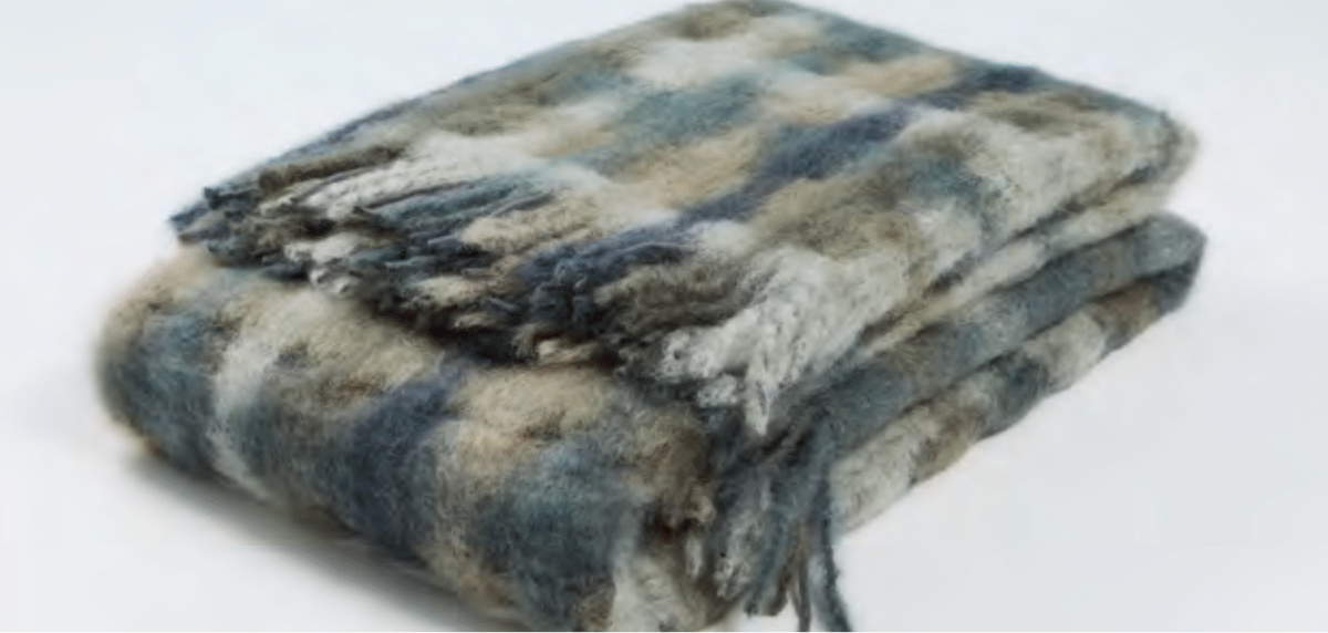 A folded, fuzzy Mia Throw by Mantas Ezcaray in shades of gray, blue, and beige with fringed edges and artisanal quality is displayed on a plain white surface.