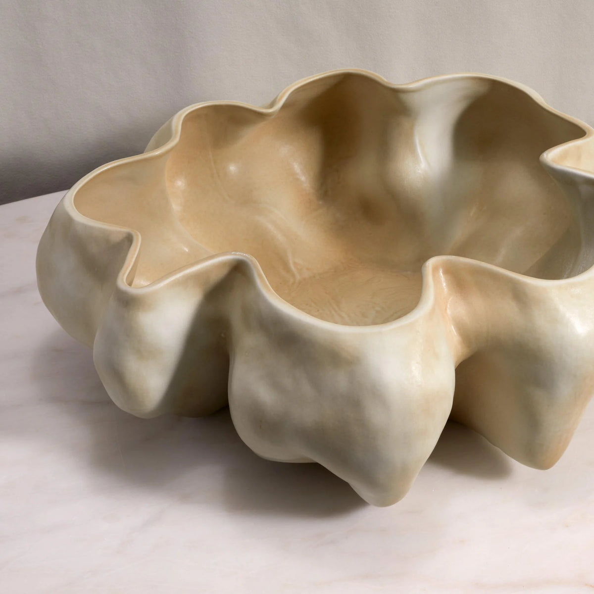 The Timna Bowl Latte by L’Objet is an off-white, hand sculpted porcelain vessel with a glossy, uneven flower-inspired interior and organic, wavy edge. It sits on a light marble surface against a neutral background.