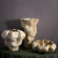 L'Objet's Timna Latte Vase Short is a sculptural porcelain vessel with organic, wavy edges in glossy off-white, displayed on a dark green surface against a plain gray backdrop.
