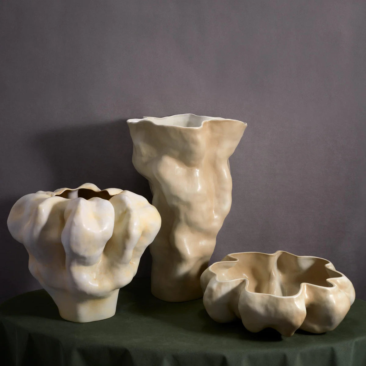 The Timna Bowl Latte by L'Objet features hand-sculpted, cream-colored ceramic bowls with unique, organic wavy forms and a glossy finish, displayed on a dark green cloth against a neutral gray background.