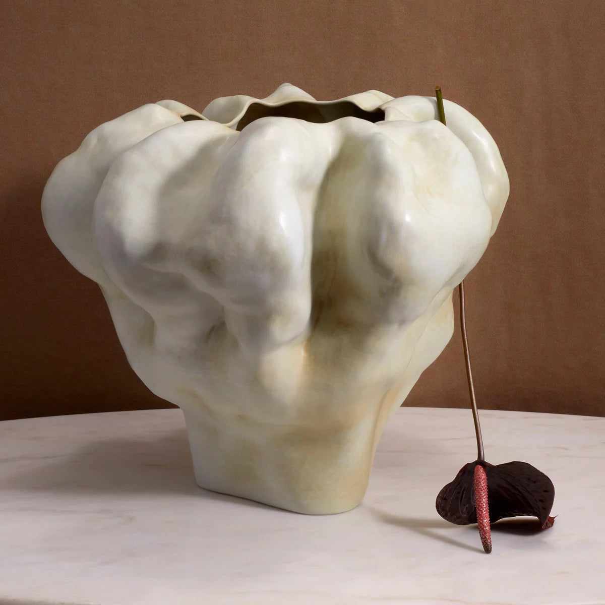 The Timna Latte Vase Short by L'Objet, a sculptural porcelain vessel with an irregular, cloud-like form, sits on a white tabletop. A dark wilted flower leans against it, echoing the stark beauty of the Arava desert on a soft brown background.