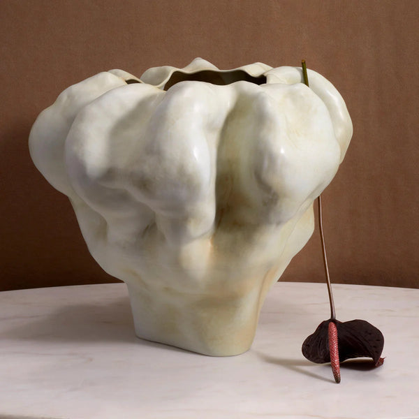 The Timna Latte Vase Short by L'Objet, a sculptural porcelain vessel with an irregular, cloud-like form, sits on a white tabletop. A dark wilted flower leans against it, echoing the stark beauty of the Arava desert on a soft brown background.