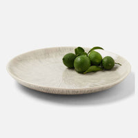 A round, textured Nolan Dinnerware Black Cream plate by Blue Pheasant holds five whole green limes and a few leaves on one side, set against a plain white background.