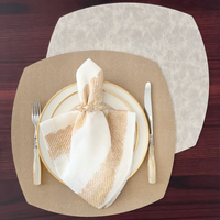 A dining table set with a beige Bodrum Stingray Elliptic Square Placemat, a white plate with a gold rim, fork on the left, knife on the right, and a white and gold napkin in a decorative ring on a dark wood surface.