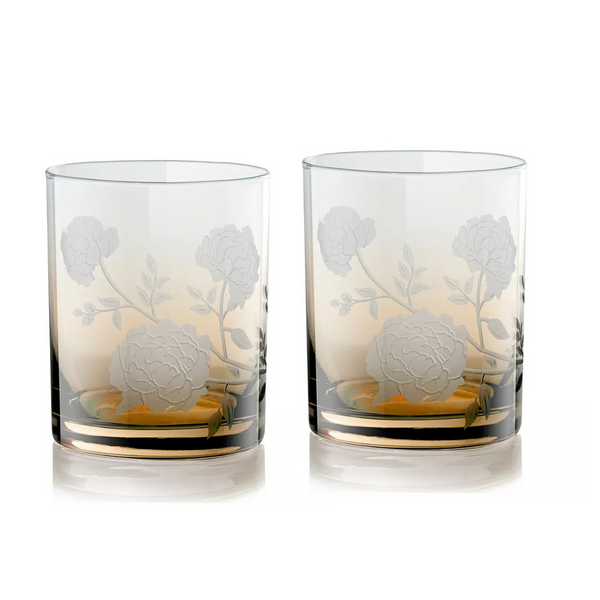 The Current Home Peony DOF Set of 2 features clear mouth-blown glass tumblers, each adorned with delicate peony flower engravings. Placed together on a white surface, they reflect light and bring elegant charm to any table setting.