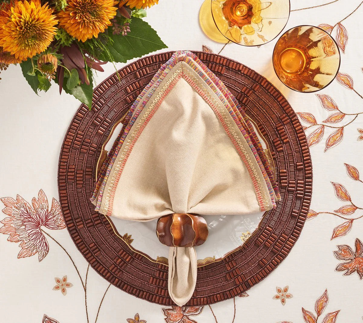 A fall-themed table setting with a woven brown placemat, cream-trim napkin held by Kim Seybert’s Twist Napkin Ring Set of 4 in Brown & Gold, floral-embroidered tablecloth, amber glassware, and a bouquet of yellow flowers.