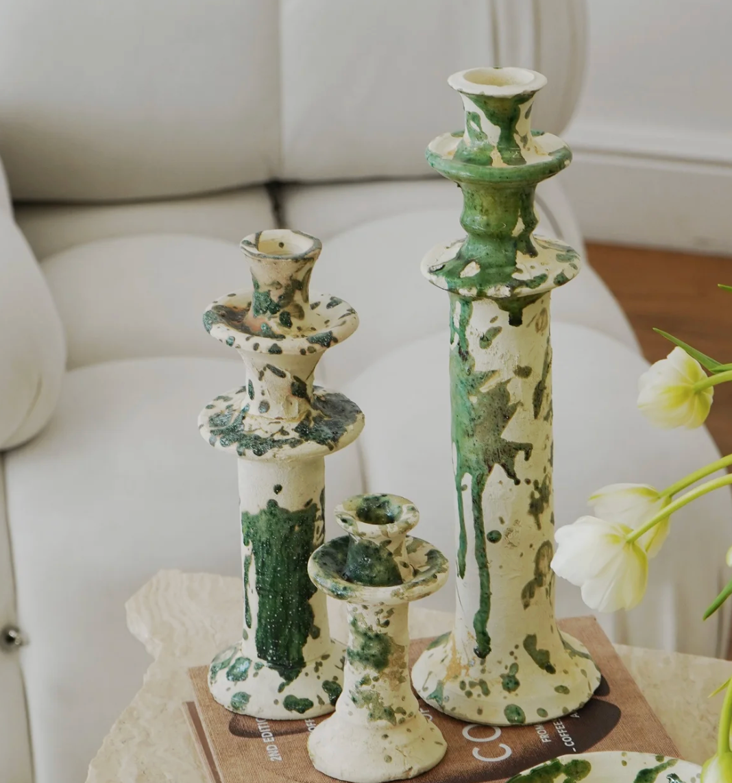 Three Vintage Splatter Candlestick Green holders by Moroccan White, with a green and white glaze, are arranged on a beige stone table beside a book. White tulips and a white couch create an elegant, artistic setting in the background.