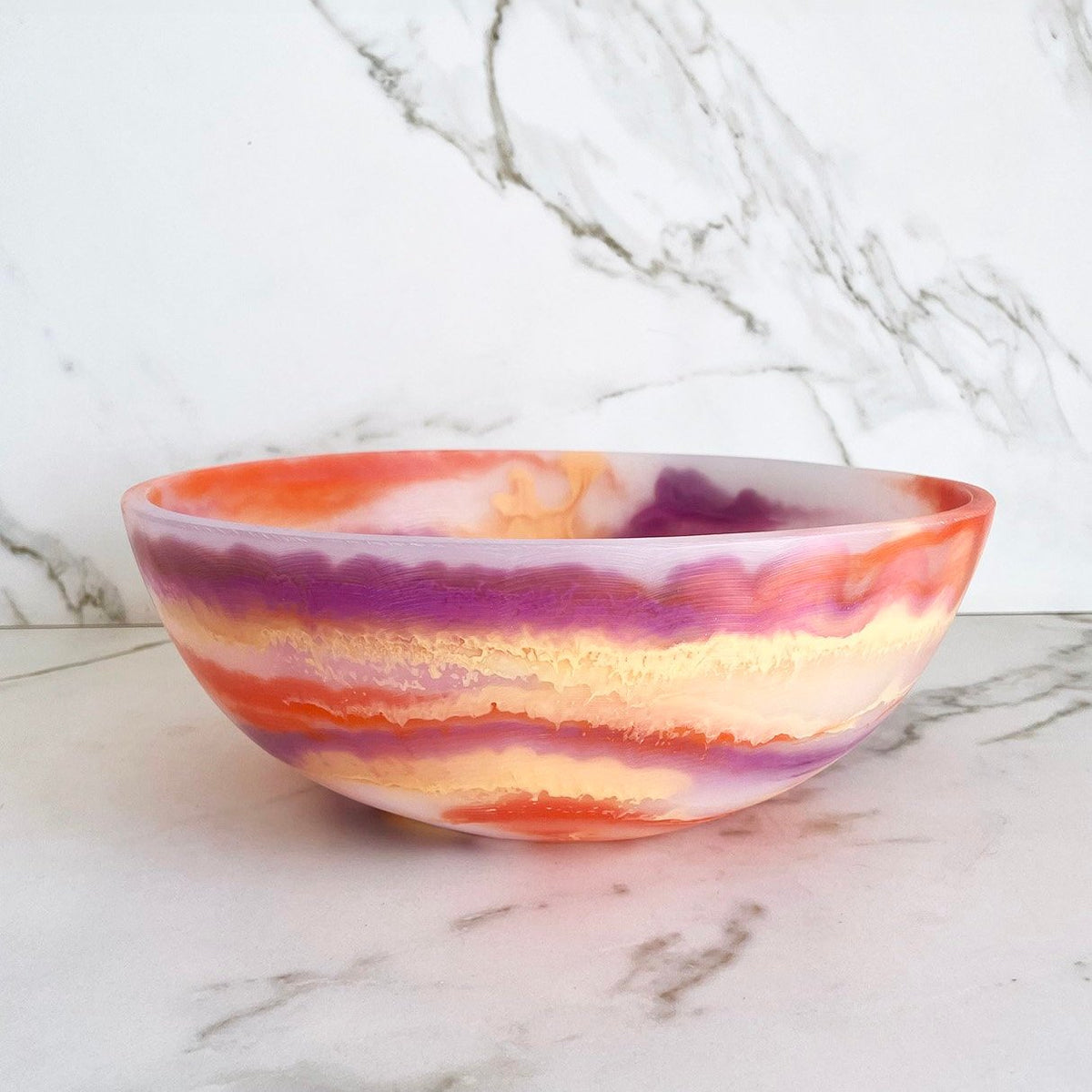 The Redondo Bowl Sherbert by CDMX / PAOLA VALLE features a marbled pattern in orange, purple, yellow, and white resin. Food safe and vibrant, it brings colorful charm to any space. Displayed on a marble surface with a marble-patterned wall behind.