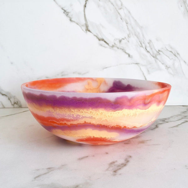 The Redondo Bowl Sherbert by CDMX / PAOLA VALLE features a marbled pattern in orange, purple, yellow, and white resin. Food safe and vibrant, it brings colorful charm to any space. Displayed on a marble surface with a marble-patterned wall behind.