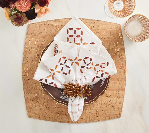 A formal table features a square textured placemat, decorative plate, and a folded Kim Seybert Malta Embroidered Napkin (Set of 4) in a beaded ring. Glassware and flowers complete the elegant setting.