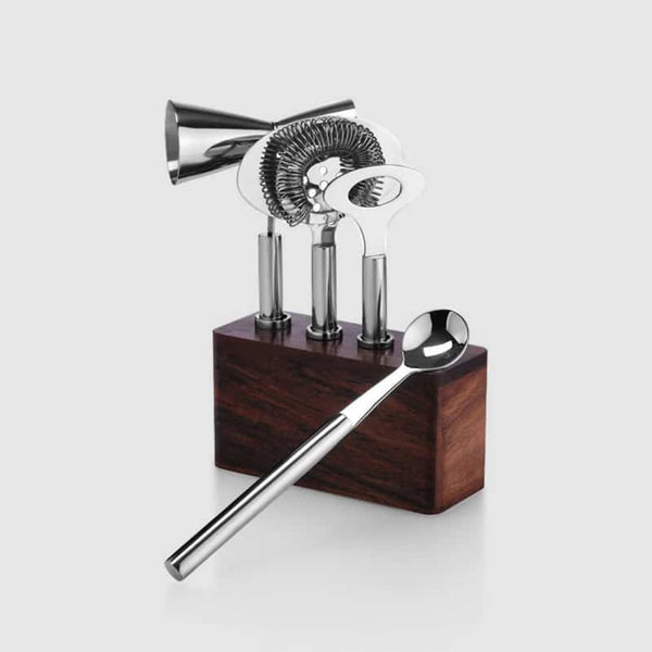 The Cosmo Bar 4 pc Set On Wood Base by Mary Jurek features polished stainless steel bar tools—a jigger, strainer, bottle opener—and a long stirring spoon, all elegantly arranged in a wooden holder for effortless cocktail crafting.