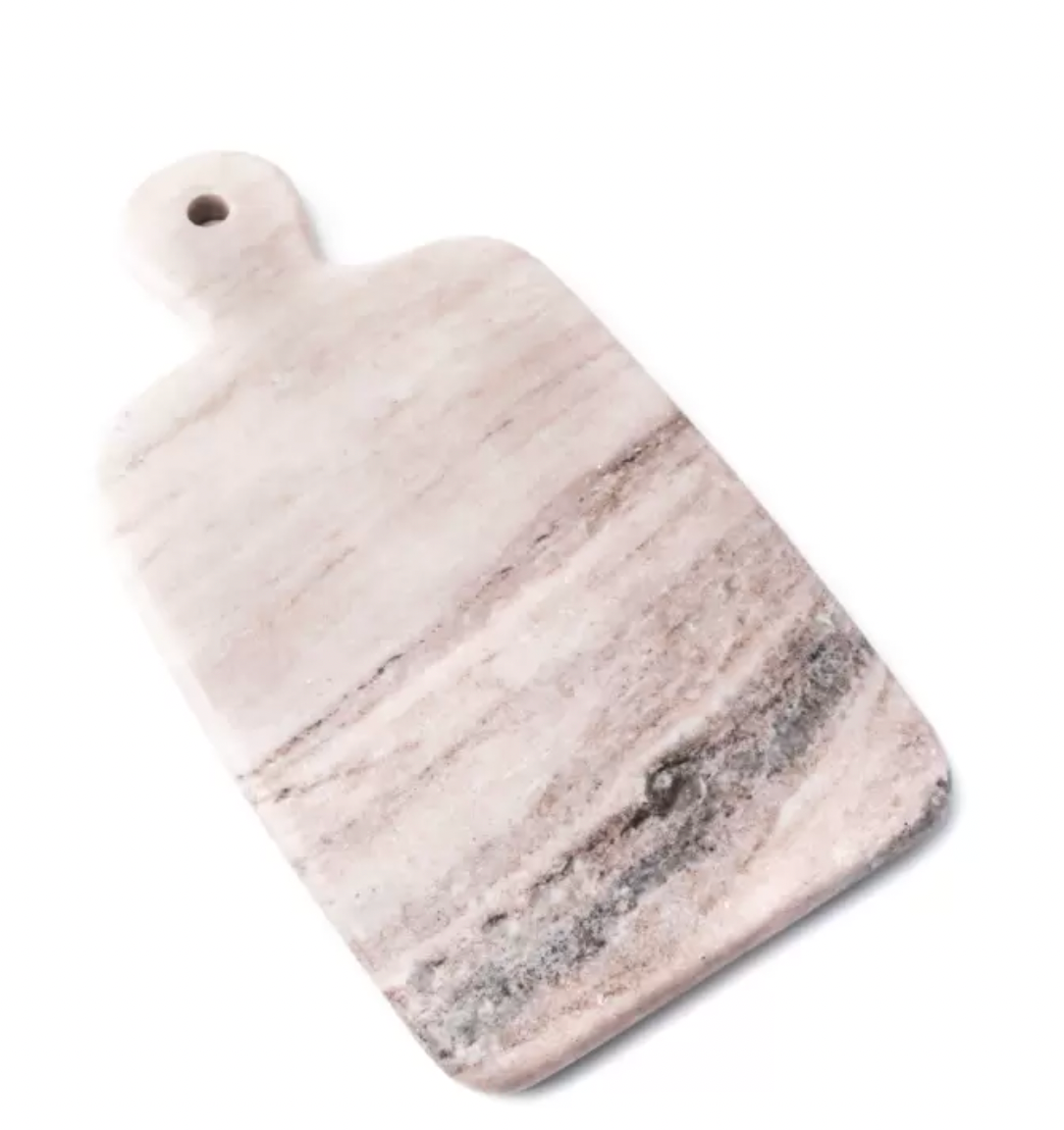 The Simon Pearce Marble Board Medium is a rectangular cutting board with rounded edges and a handle with a small hole. Displayed on a white background, its marble surface features light pinkish and gray natural veining.