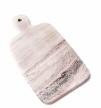 The Simon Pearce Marble Board Medium is a rectangular cutting board with rounded edges and a handle with a small hole. Displayed on a white background, its marble surface features light pinkish and gray natural veining.