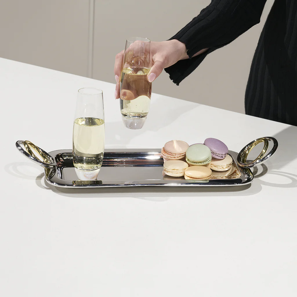 A person picks up a glass of sparkling wine from the Anna New York Ring Rectangle Serving Tray, which features gold handles and also holds another glass and colorful macarons, all displayed on a white surface.