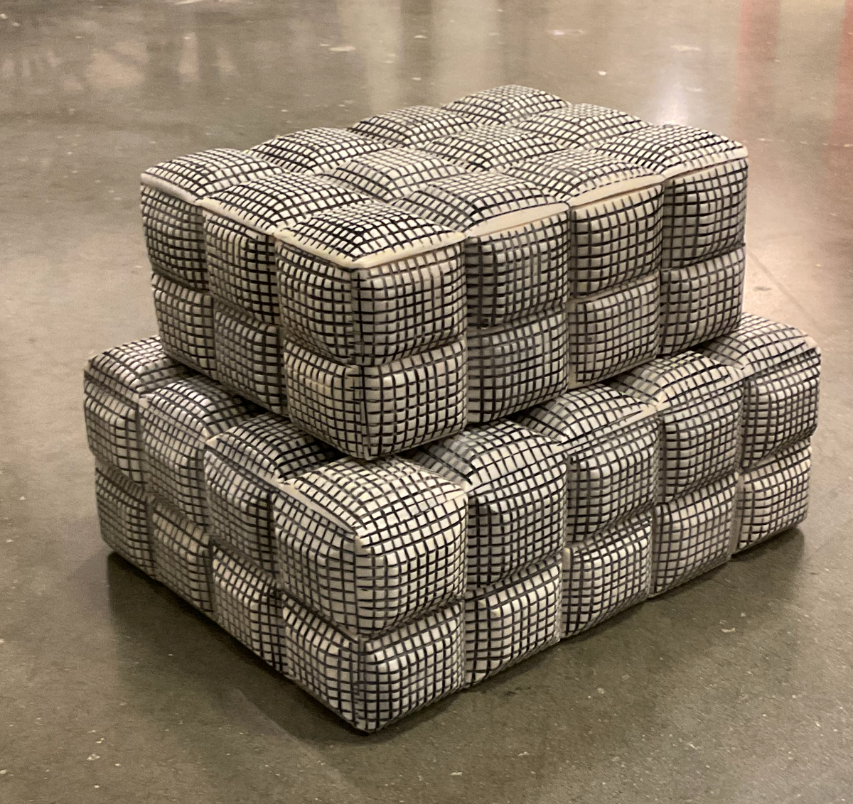 Two stacked Cubie Box poufs by A Sanoma Inc with a black grid pattern on cream sit on a concrete floor, adding an edgy decorative accent to your home.