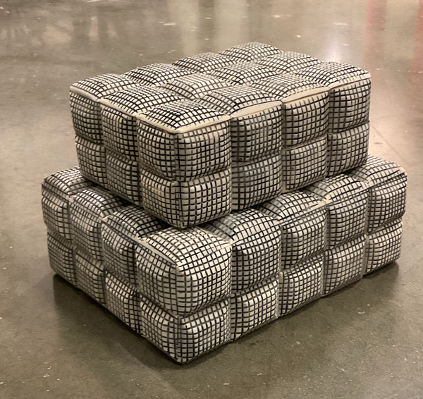 Two stacked Cubie Box poufs by A Sanoma Inc with a black grid pattern on cream sit on a concrete floor, adding an edgy decorative accent to your home.