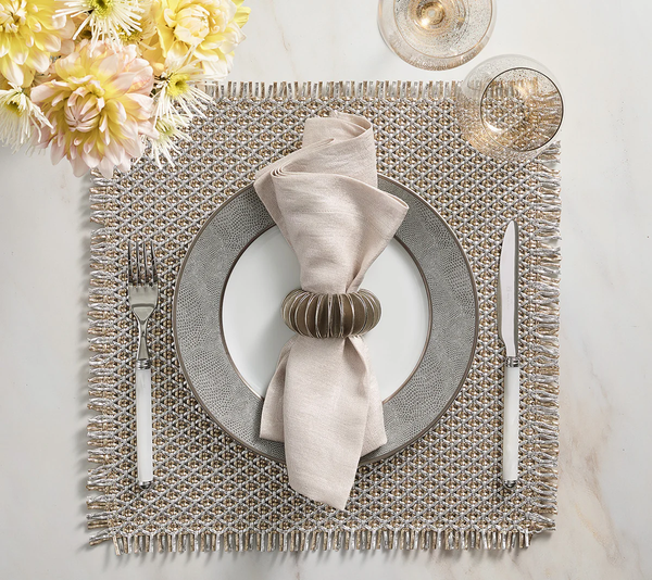 A refined table setting featuring a textured placemat, ornate plate, neatly folded beige napkin in the Kim Seybert Demilune Napkin Ring Silver Set of 4, with silverware, wine glass, and pale yellow flowers on a white surface.