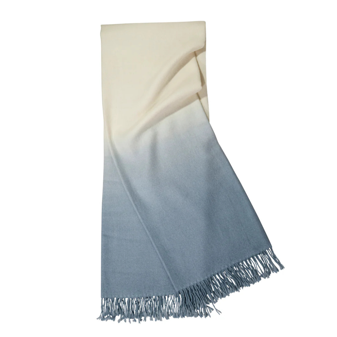 The Johanna Howard Dip Dye Throw features a gradient ombré design that transitions from cream to light blue, finished with stylish fringed edges.