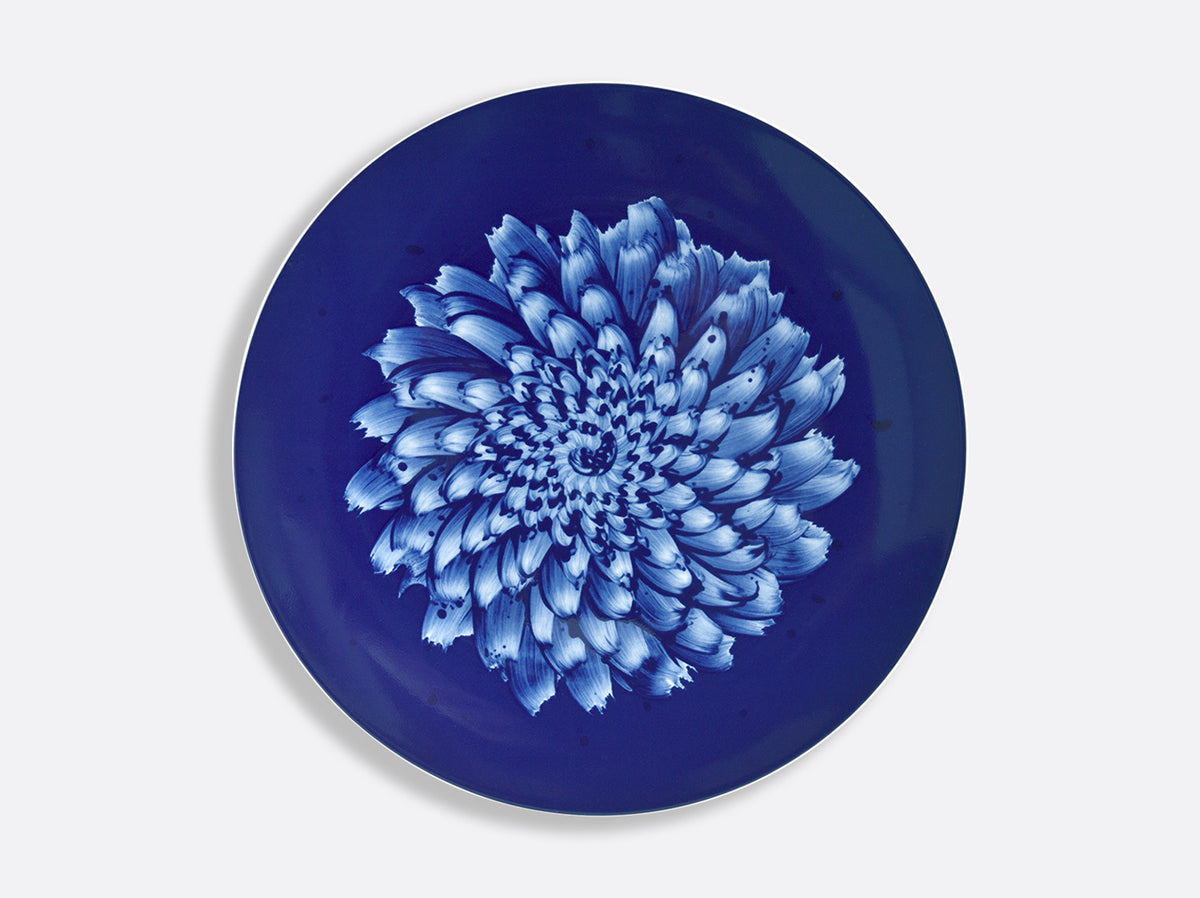 The In Bloom Dinnerware by Bernardaud features a round plate with a deep blue background and an intricate white and light blue chrysanthemum-inspired floral design at the center, reflecting the artistry of Zemer Peled's porcelain collection.