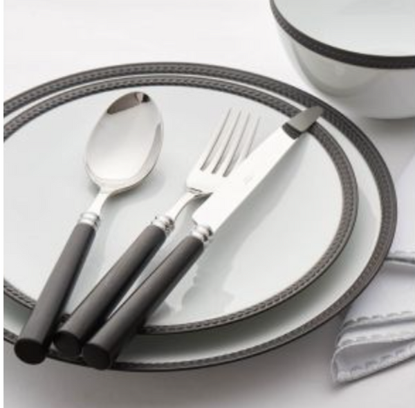 A plate and bowl with a dark rim are set with Capdeco Marat Wood Flatware—fork, knife, and spoon featuring black handles—arranged on a white napkin atop a white surface.