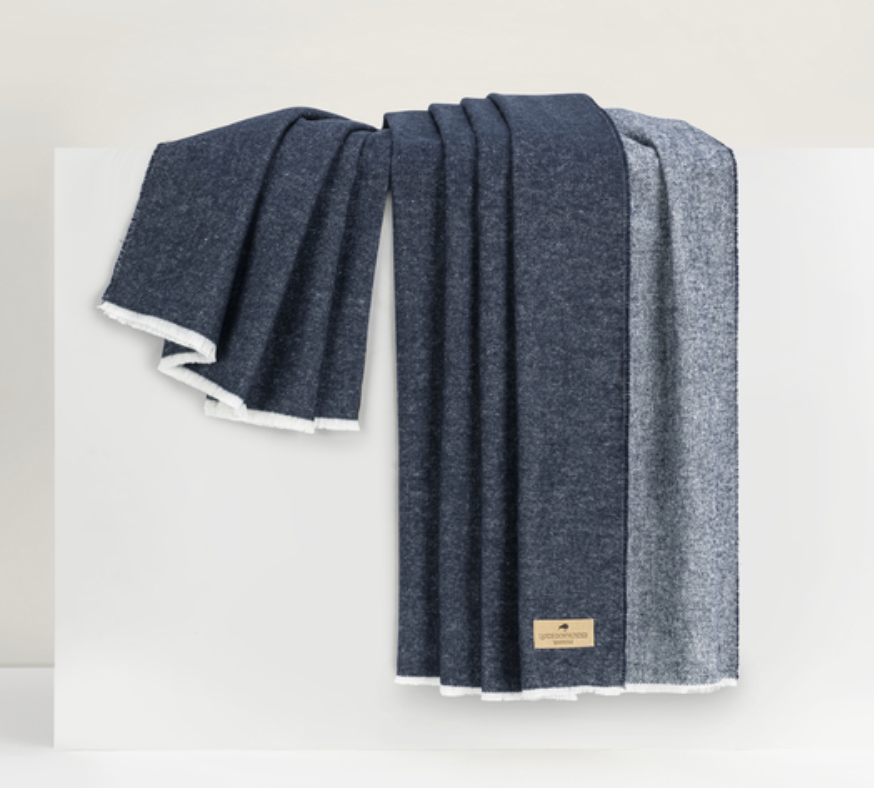 The Aspen Reversible Throw by Lands Downunder, featuring dark blue and gray hues with a soft white lining and a tan fabric label, is elegantly draped over a white surface for a luxurious look.