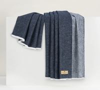 The Aspen Reversible Throw by Lands Downunder, featuring dark blue and gray hues with a soft white lining and a tan fabric label, is elegantly draped over a white surface for a luxurious look.