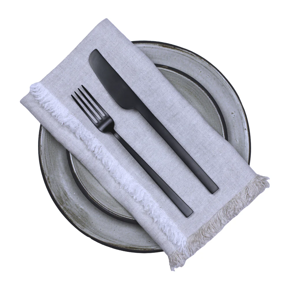 Two ceramic plates are stacked with a Hogar Frayed Edge Linen Napkin by Linen Casa on top, and set with a black fork and knife. The white background complements the refined yet relaxed style.