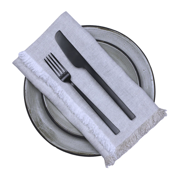 Two ceramic plates are stacked with a Hogar Frayed Edge Linen Napkin by Linen Casa on top, and set with a black fork and knife. The white background complements the refined yet relaxed style.