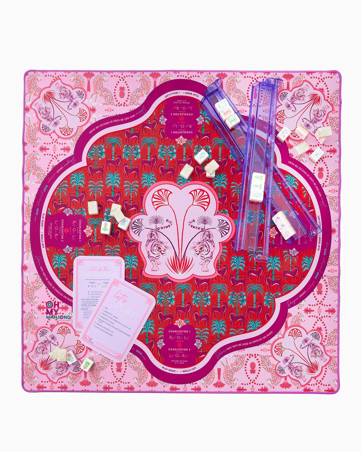 Oh My Mahjong’s Mahjong Mats feature a pink, red, and purple neoprene game mat with decorative motifs and symmetrical patterns, plus rectangular cards, sliding rulers, and small wrapped tokens for a colorful gaming experience.