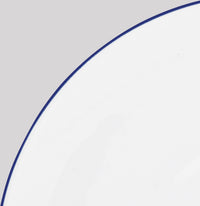 A close-up shows the Raynaud Monceau Bleu Outremer Dinnerware—a white ceramic plate with a thin ultramarine blue rim—set against a light gray background, with only part of the plate visible.